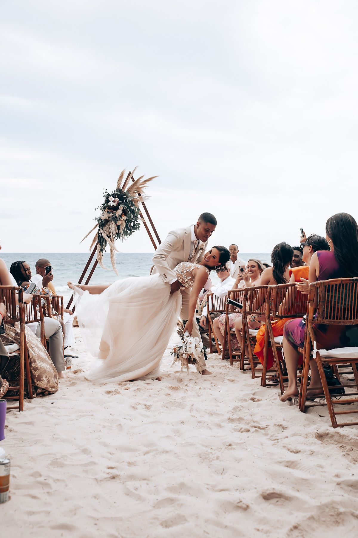Wedding guests seated on wooden chairs on sandy beach watching ceremony under boho-style arch.