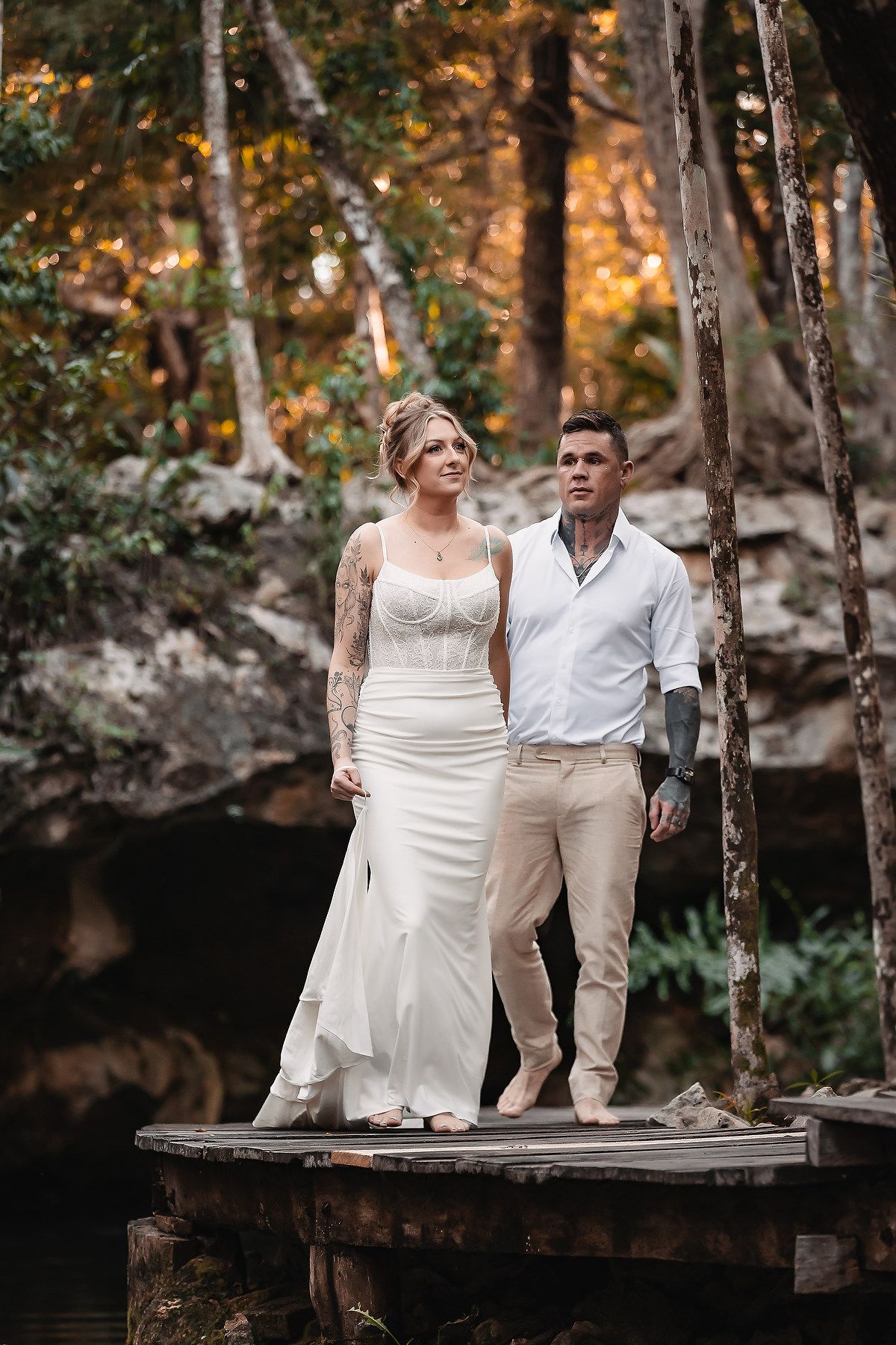 Tattooed bride and groom stand on wooden bridge surrounded by lush forest in romantic outdoor wedding portrait.