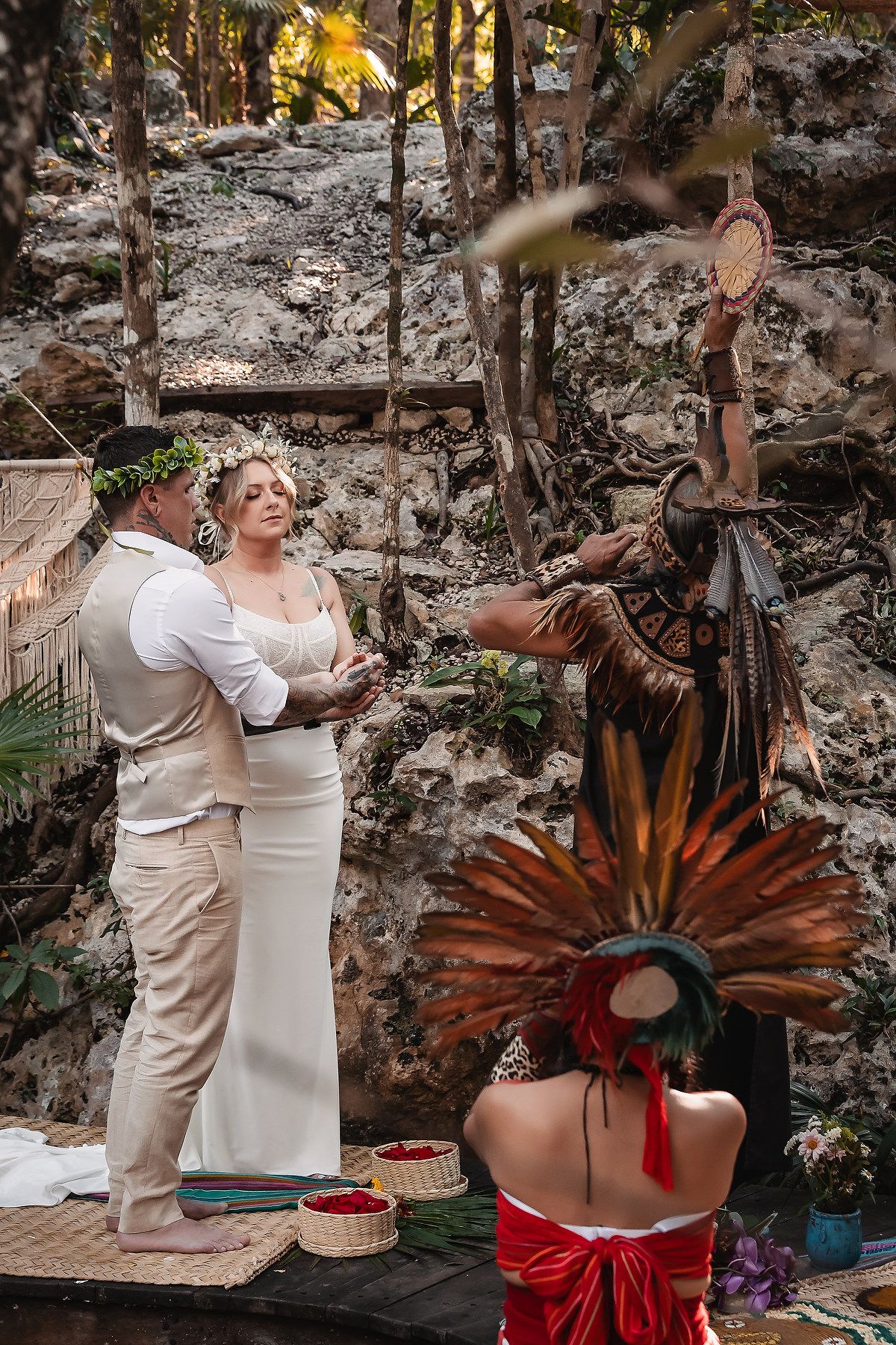 Bride and groom hold hands during Mayan ceremony in jungle with shaman in feathered headdress officiating.