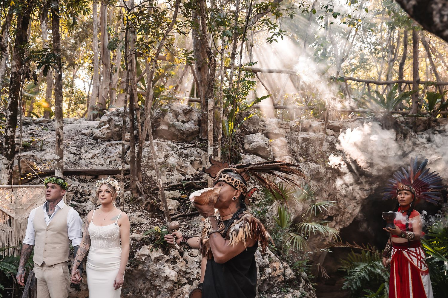 Couple in wedding attire with Mayan shaman officiating ceremony in lush jungle ruins with dramatic sunbeams.