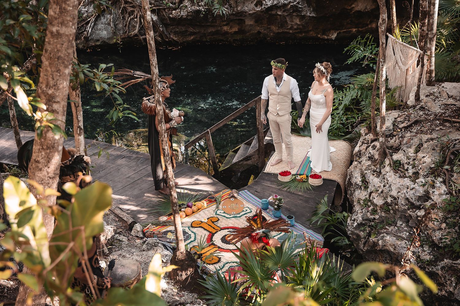Couple in wedding attire standing by a cenote surrounded by lush jungle and colorful floral ground art.