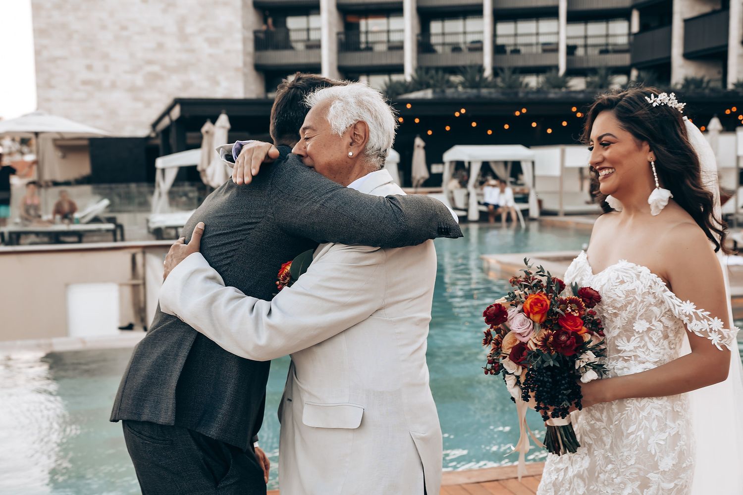 Wedding couple embracing by a poolside with decorative red floral arrangements.