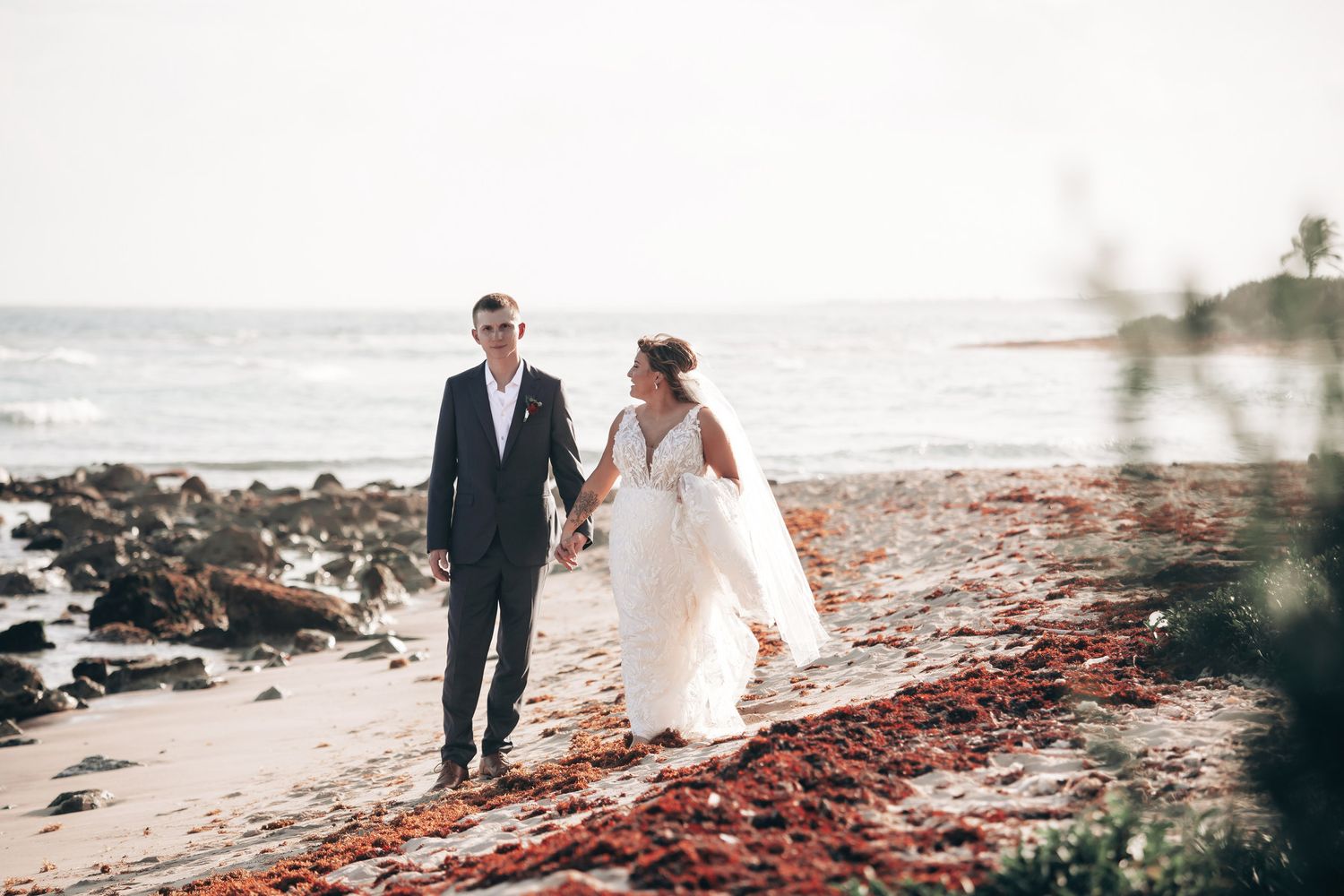A bride and groom walk hand in hand along a scenic beach with red seaweed and ocean waves behind them.