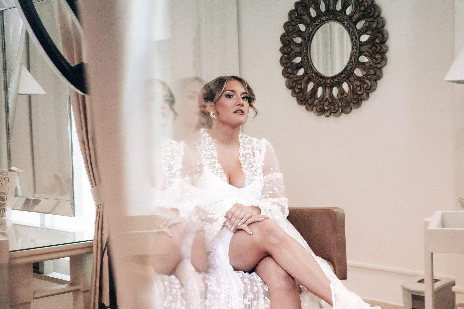 Bride in white lace robe sitting elegantly in chair before mirror in bright dressing room.