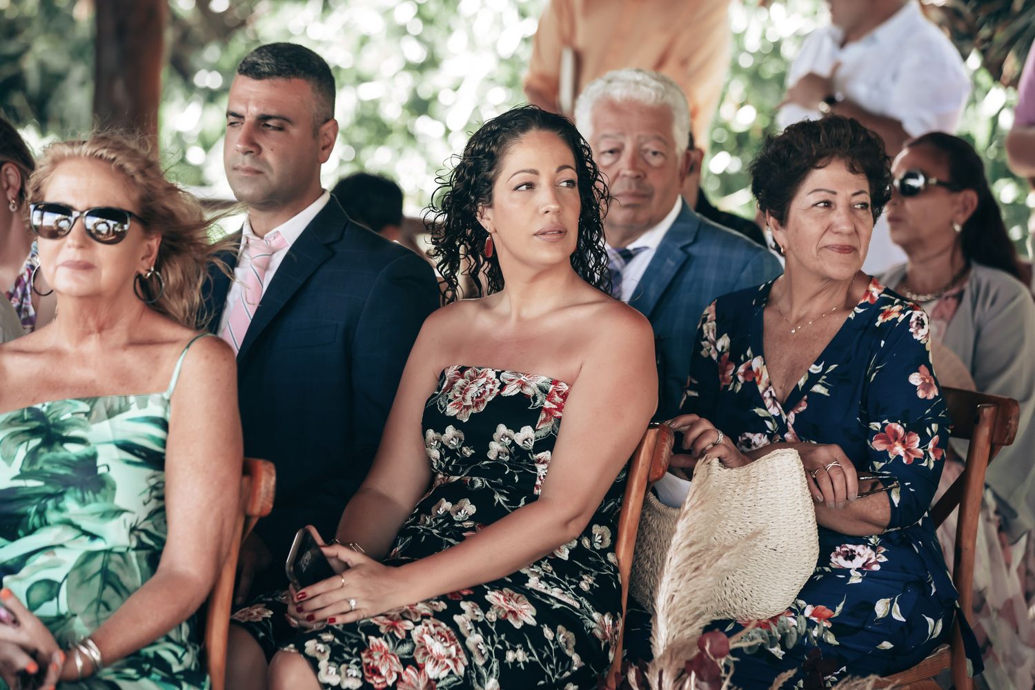 Wedding guests seated outdoors watching ceremony in floral attire on sunny day.