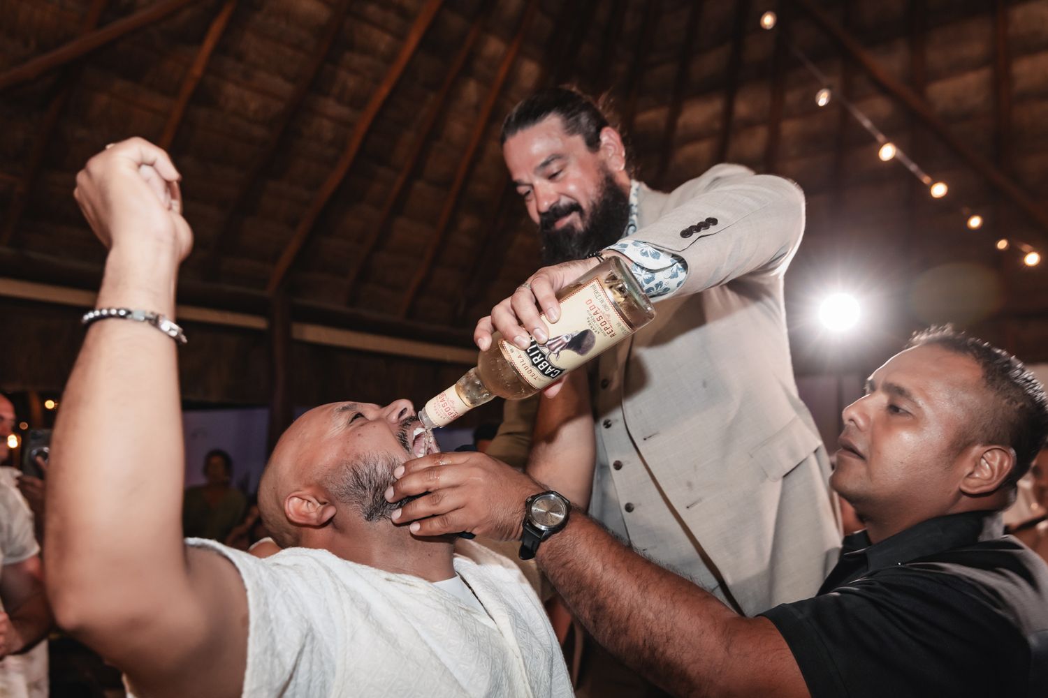 Man drinks from bottle poured by another man in beige suit at lively indoor party celebration.
