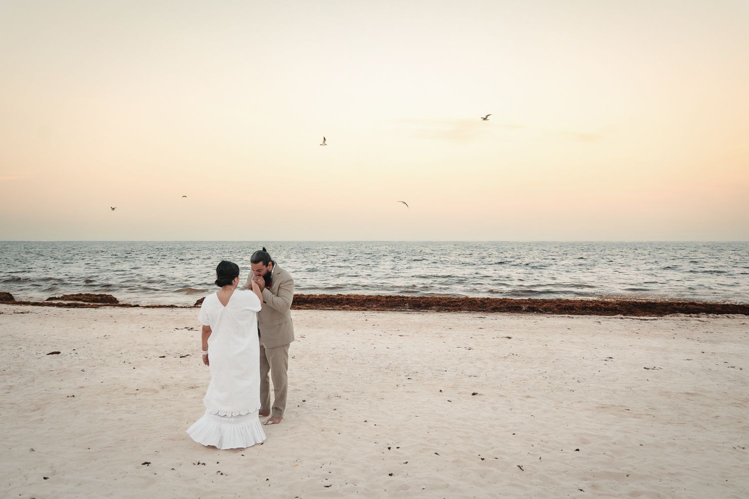 Bride and groom stand together on a sandy beach at sunset, with birds flying over the calm ocean horizon.