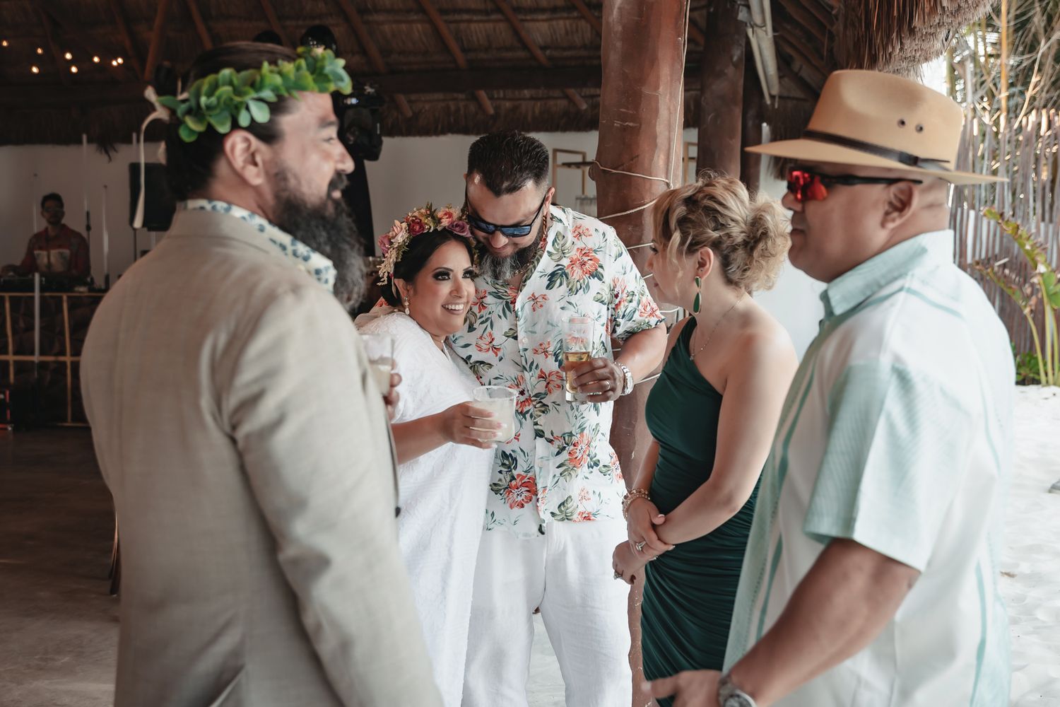 Tropical beach wedding guests in floral attire and leis socializing under a thatched roof venue.