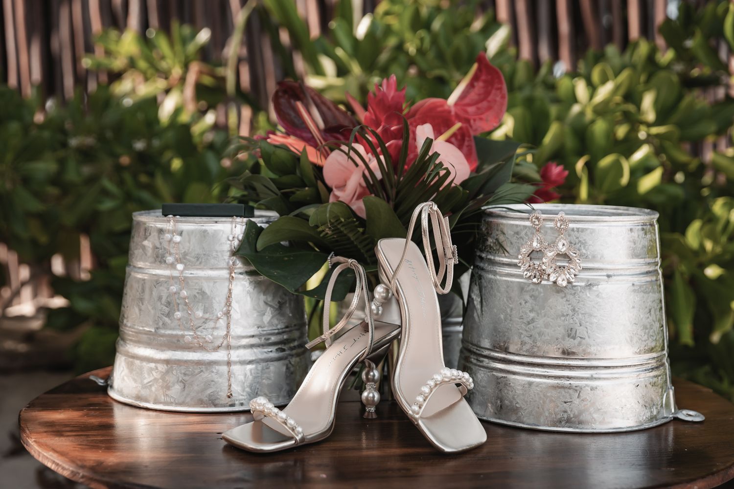 Elegant bridal heels with pearl accents displayed between rustic galvanized buckets holding tropical floral arrangements.