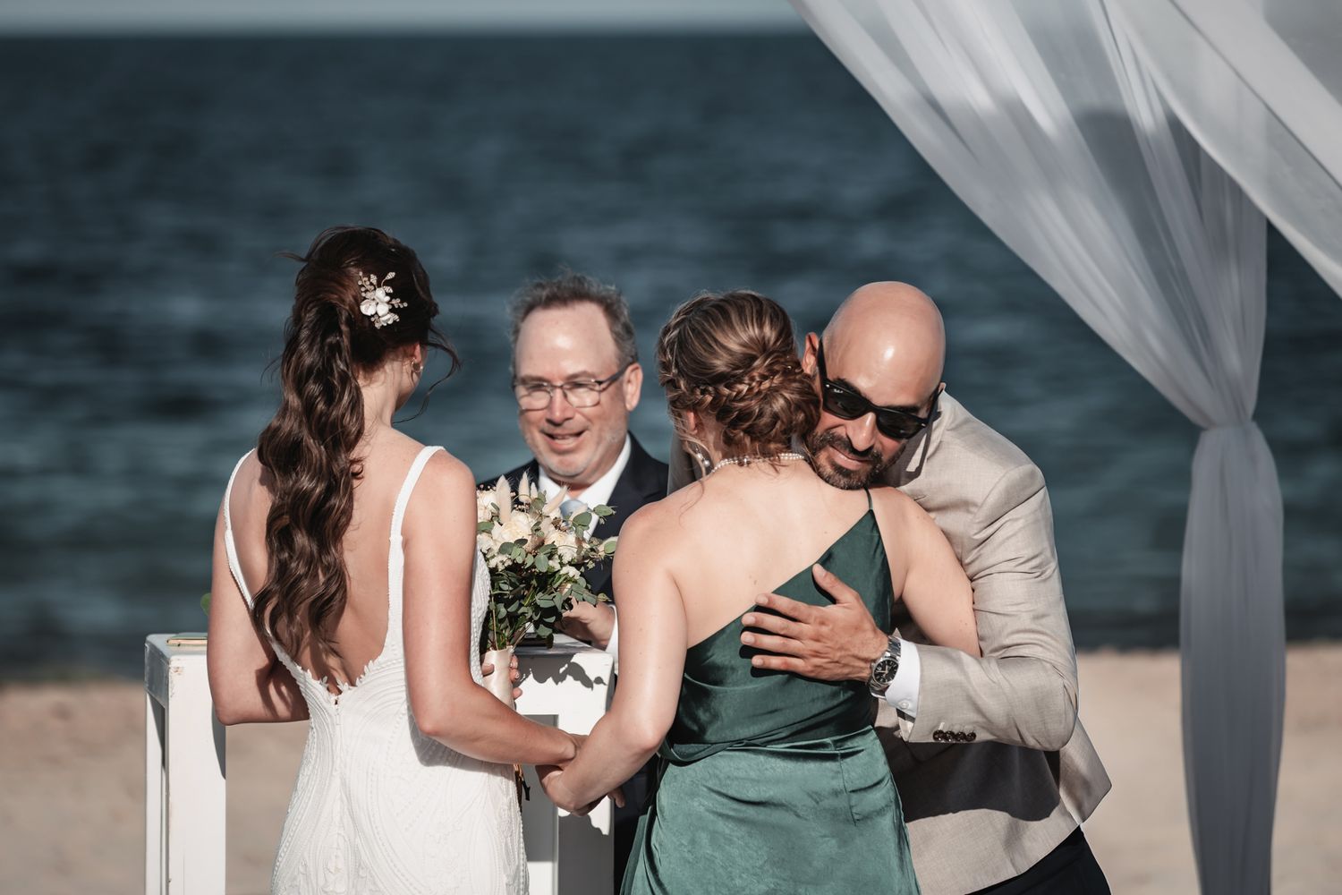 Bride and groom embrace guests during a beachside wedding ceremony with ocean backdrop.