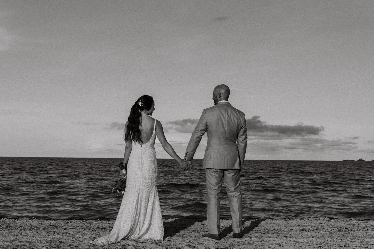 Black and white photo of a bride and groom holding hands while standing on a beach at sunset.
