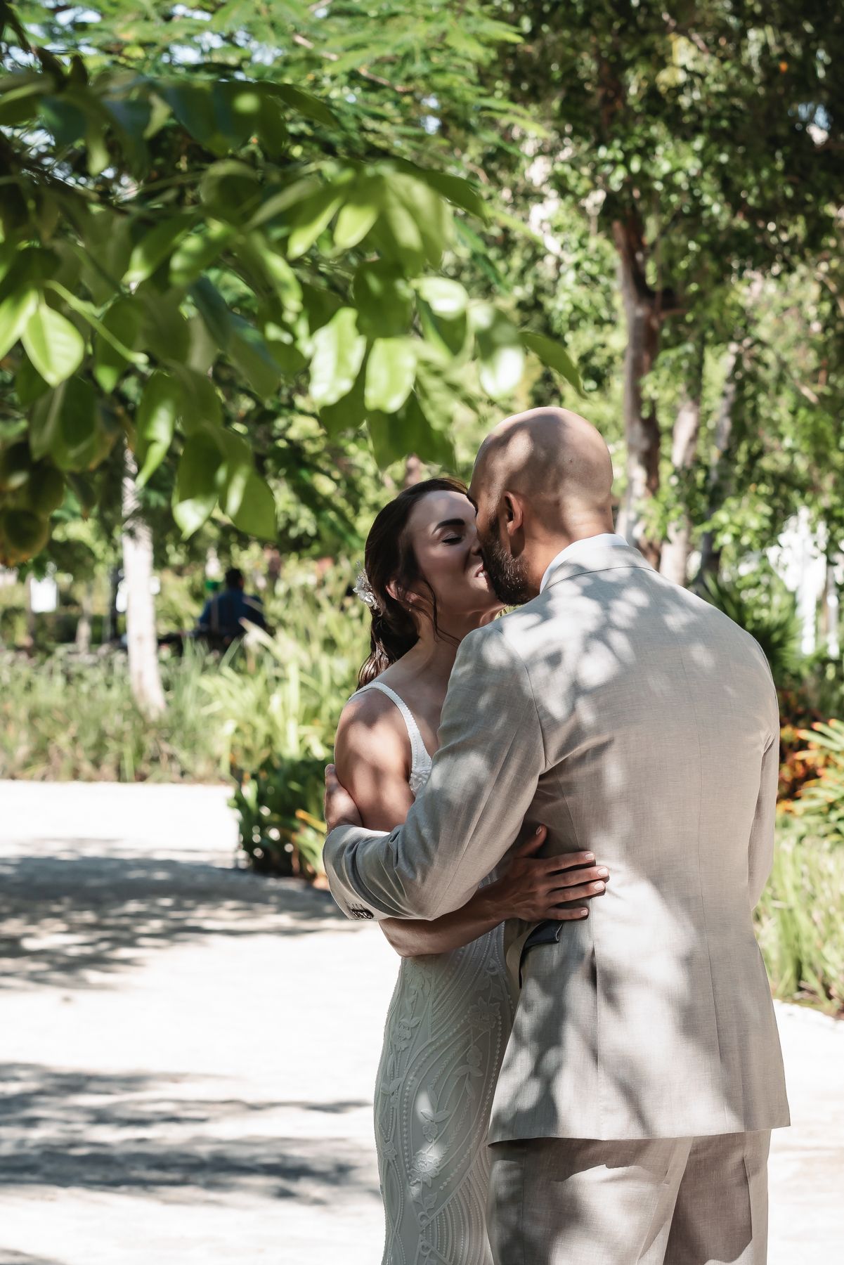 A bride and groom share a romantic kiss surrounded by lush green trees in an outdoor garden setting.