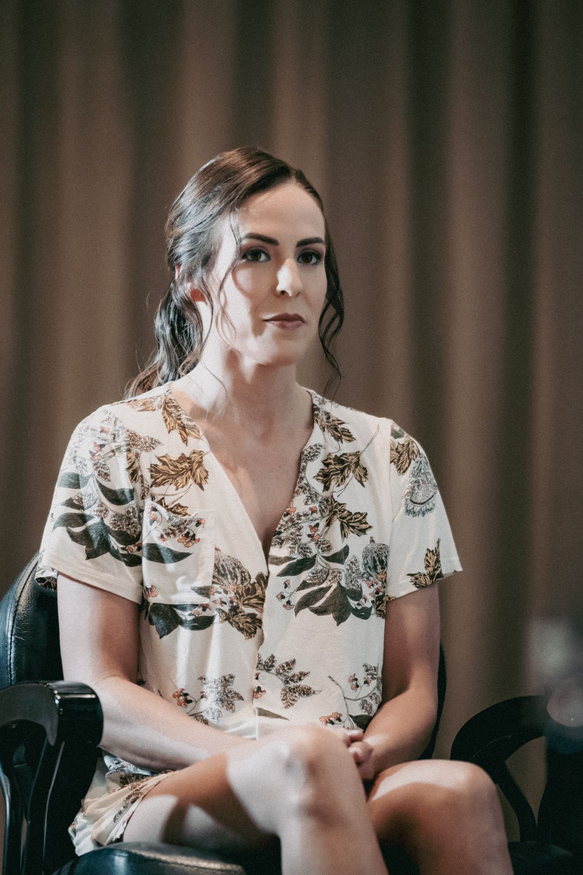 Woman in floral dress sitting in chair against curtain backdrop, looking thoughtful.