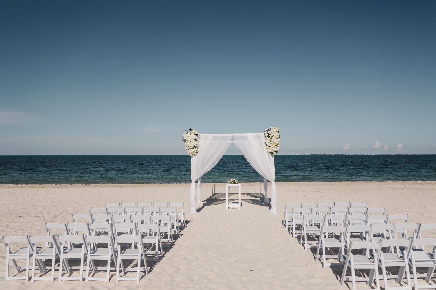 Beach wedding setup with white chairs, floral arch, and ocean backdrop on a sunny day.