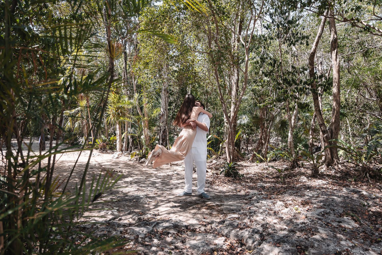 Couple embracing on a sunlit forest path surrounded by lush tropical trees and dappled light.