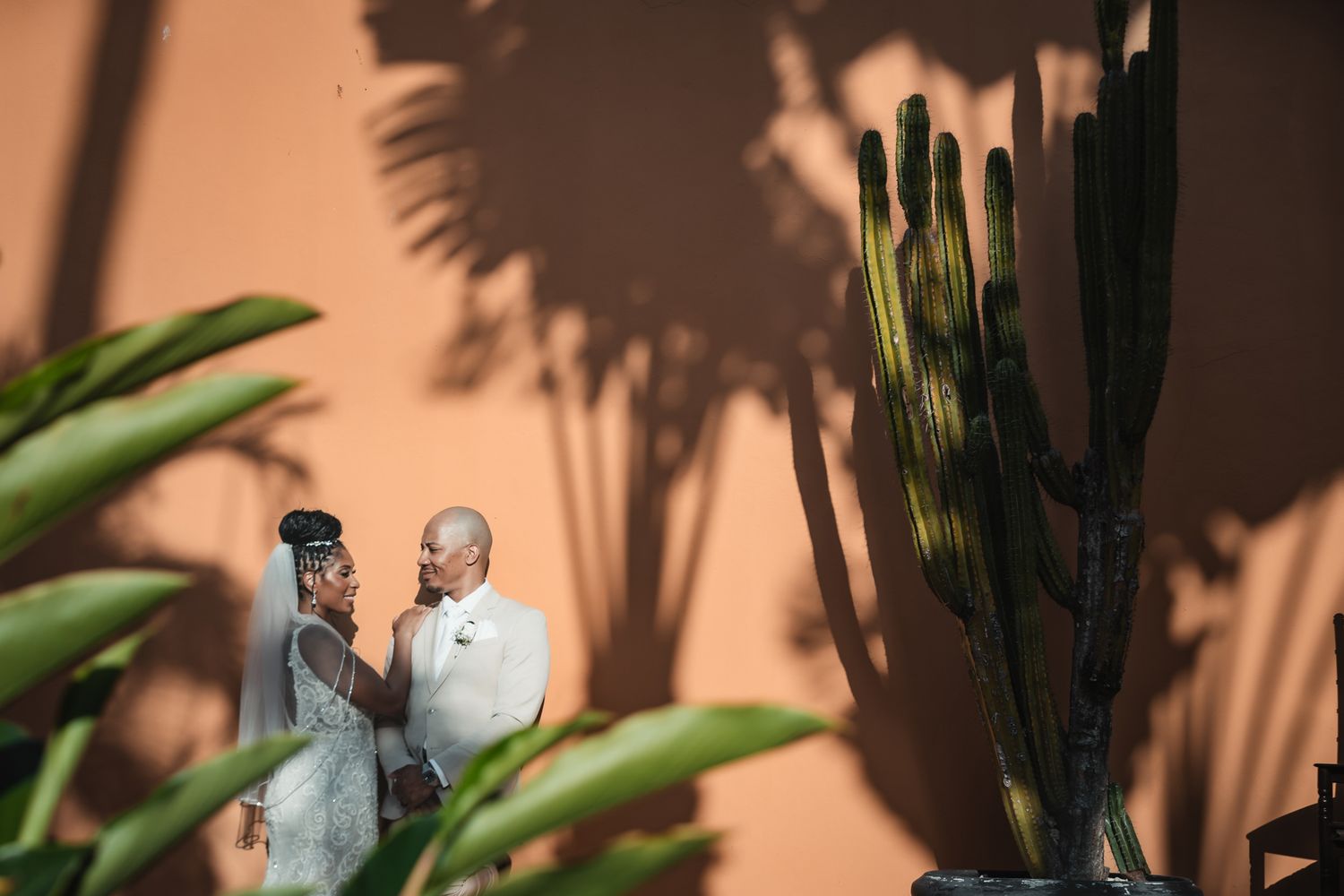 Couple shares a romantic moment by a terracotta wall with cacti and tropical plants in a desert wedding setting.