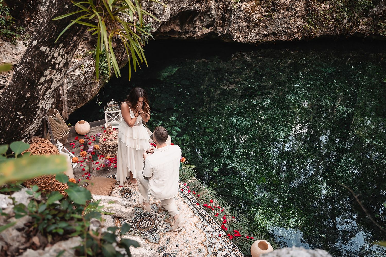 Man proposing to woman on a romantic setup beside a lush green cenote in a scenic outdoor location.