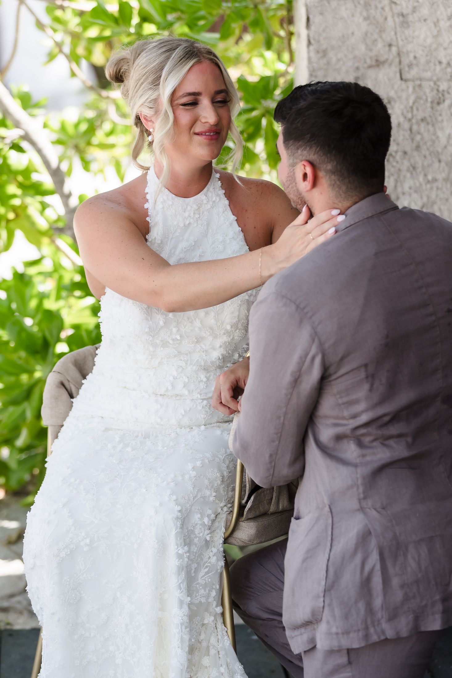 Bride in white lace gown tenderly touches groom's face during outdoor wedding ceremony with green foliage backdrop.