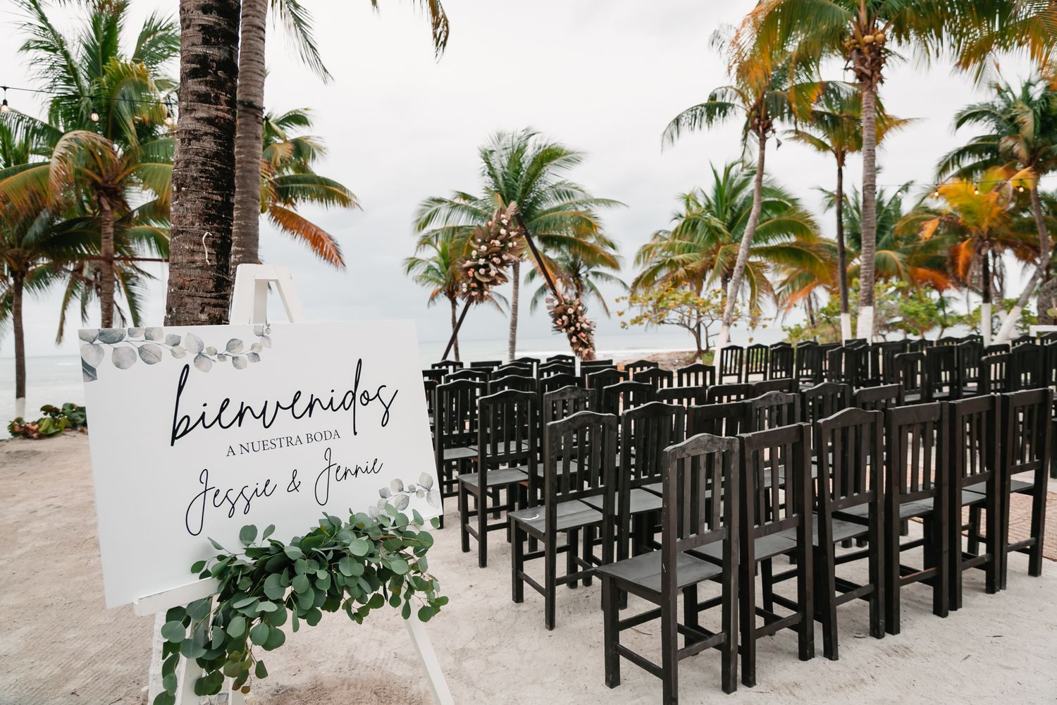 Outdoor beach wedding ceremony setup with black chairs and a bienvenidos welcome sign surrounded by palm trees.