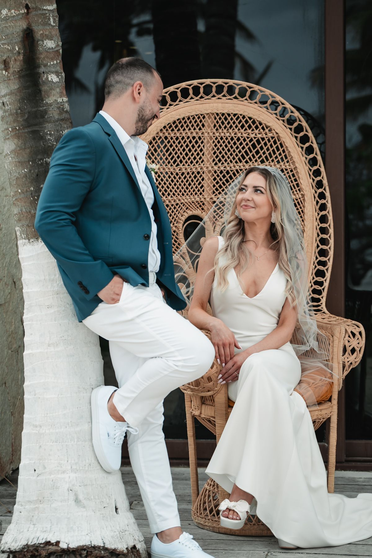 Bride in white gown seated in a rattan peacock chair beside groom in navy blazer at tropical venue.