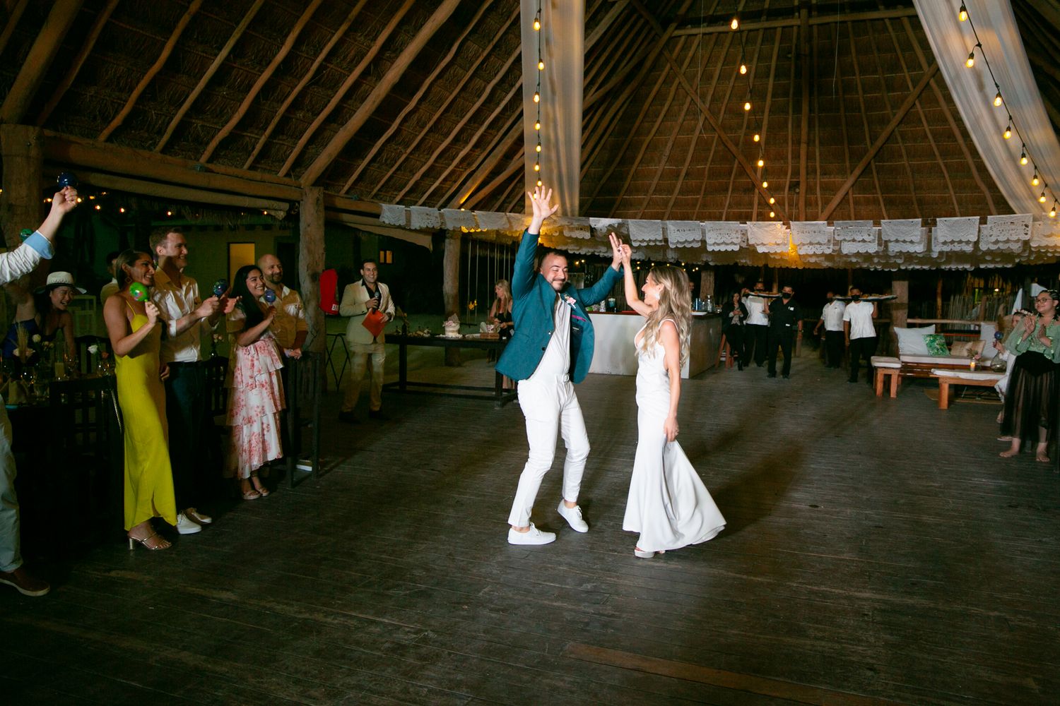 Groom in teal jacket spins bride in white gown on dance floor at tropical wedding reception.