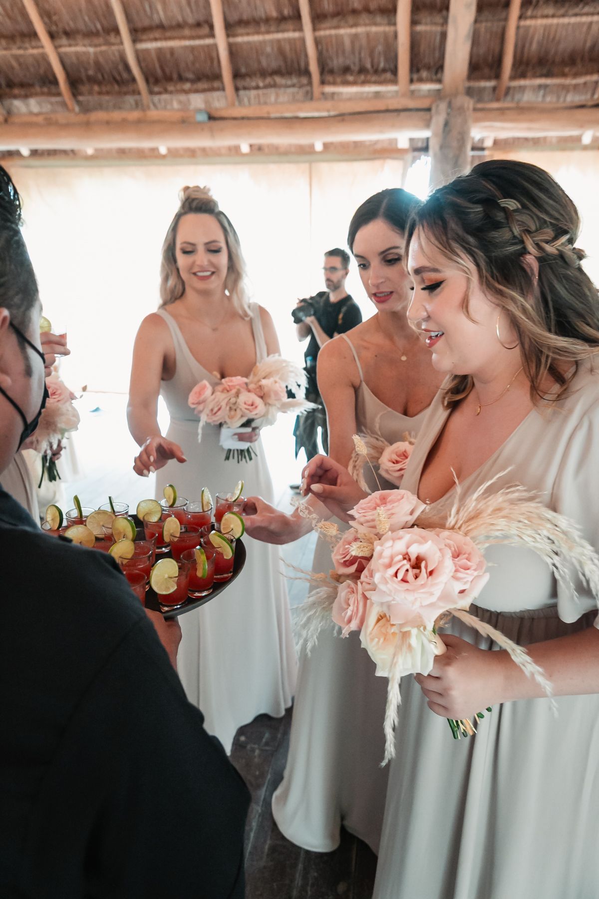 Bridesmaids in sage green dresses sample appetizers while holding pink floral bouquets at a beach wedding reception.