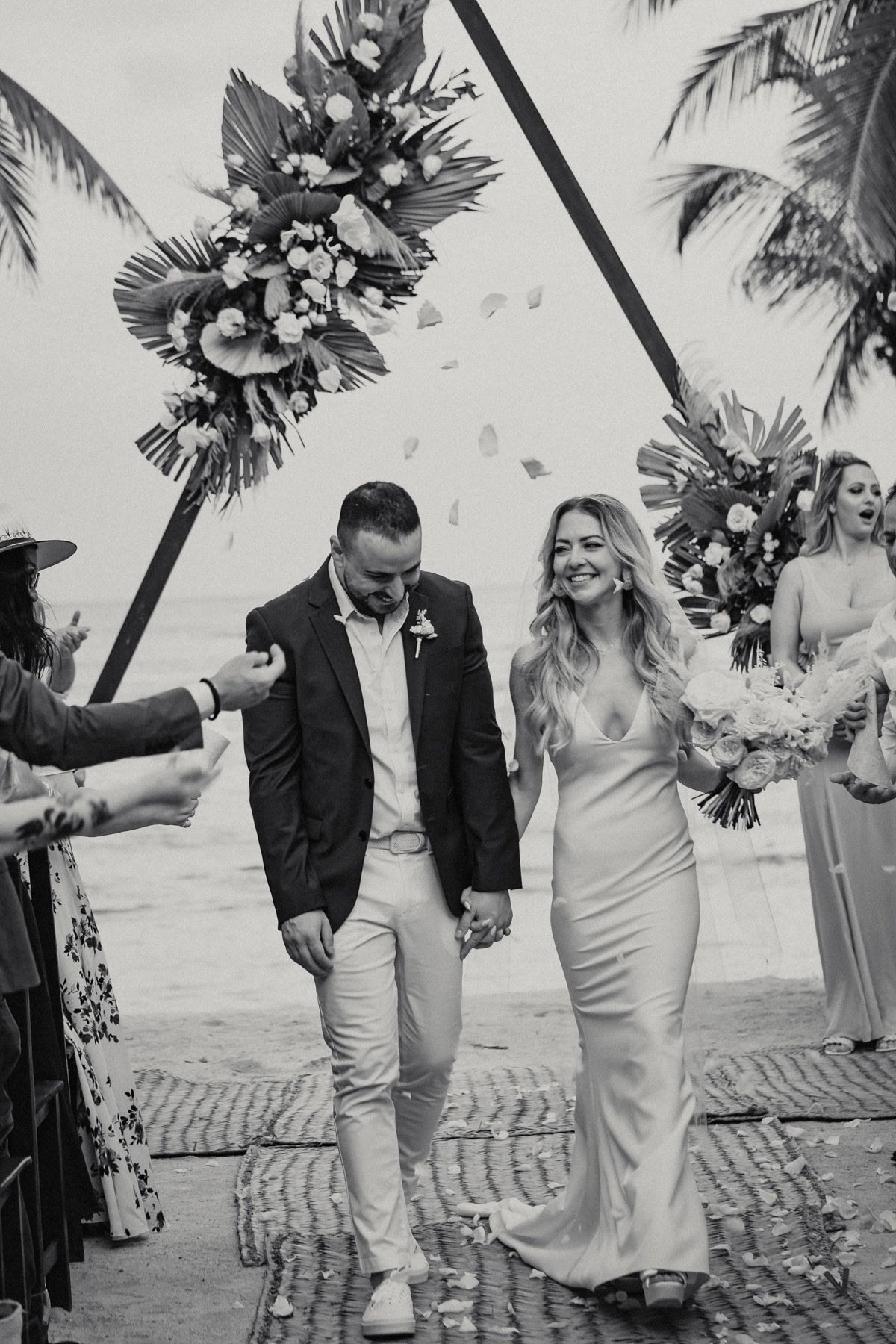 Newlyweds walk hand in hand under tropical floral arch on beach after ceremony in black and white.