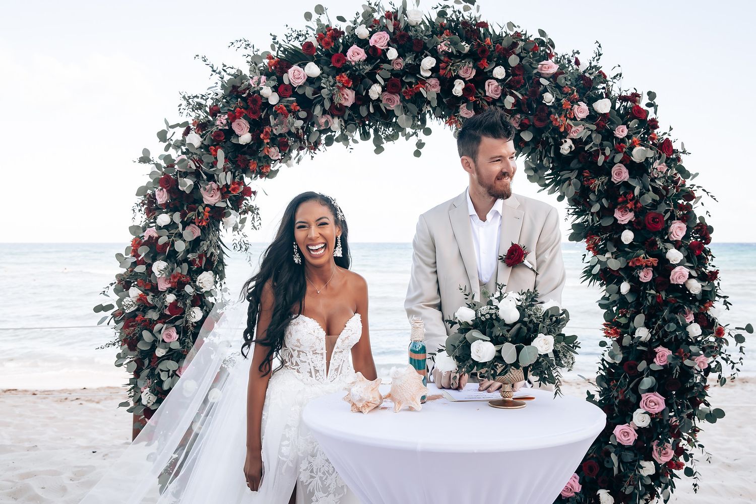 Floral wedding arch with pink and white roses frames a beachside ceremony.