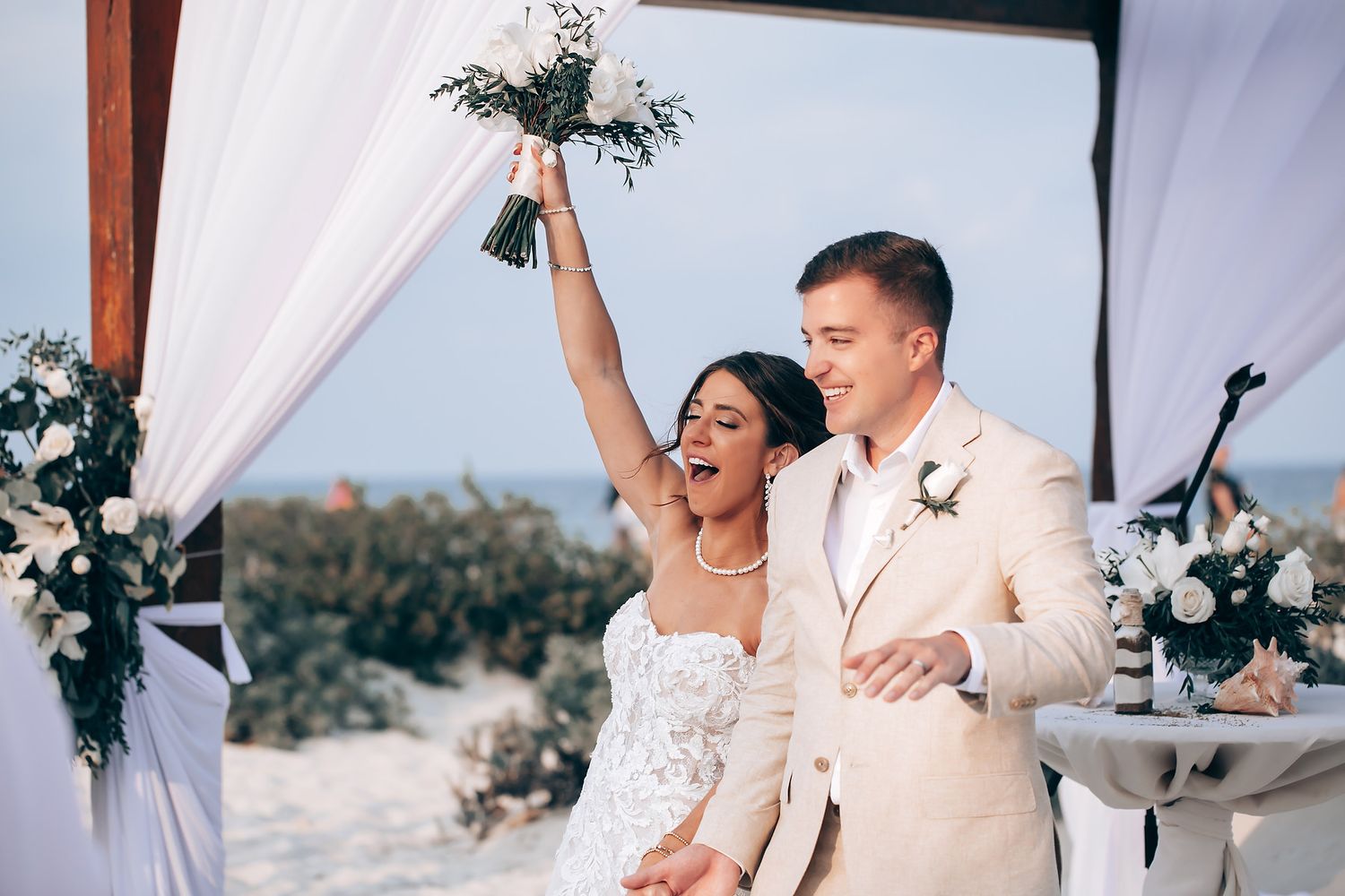 A romantic beach wedding ceremony setup with white drapes and floral arrangements against an ocean backdrop.