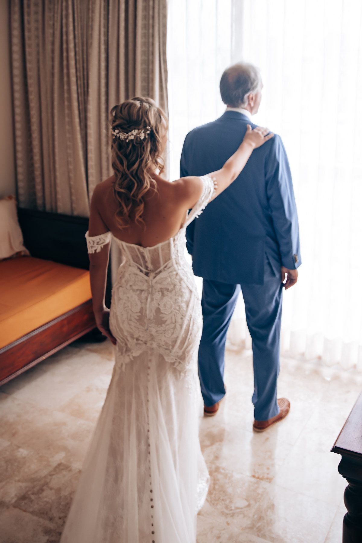 A bride in a fitted lace gown touches the shoulder of someone in a navy suit before their wedding ceremony.