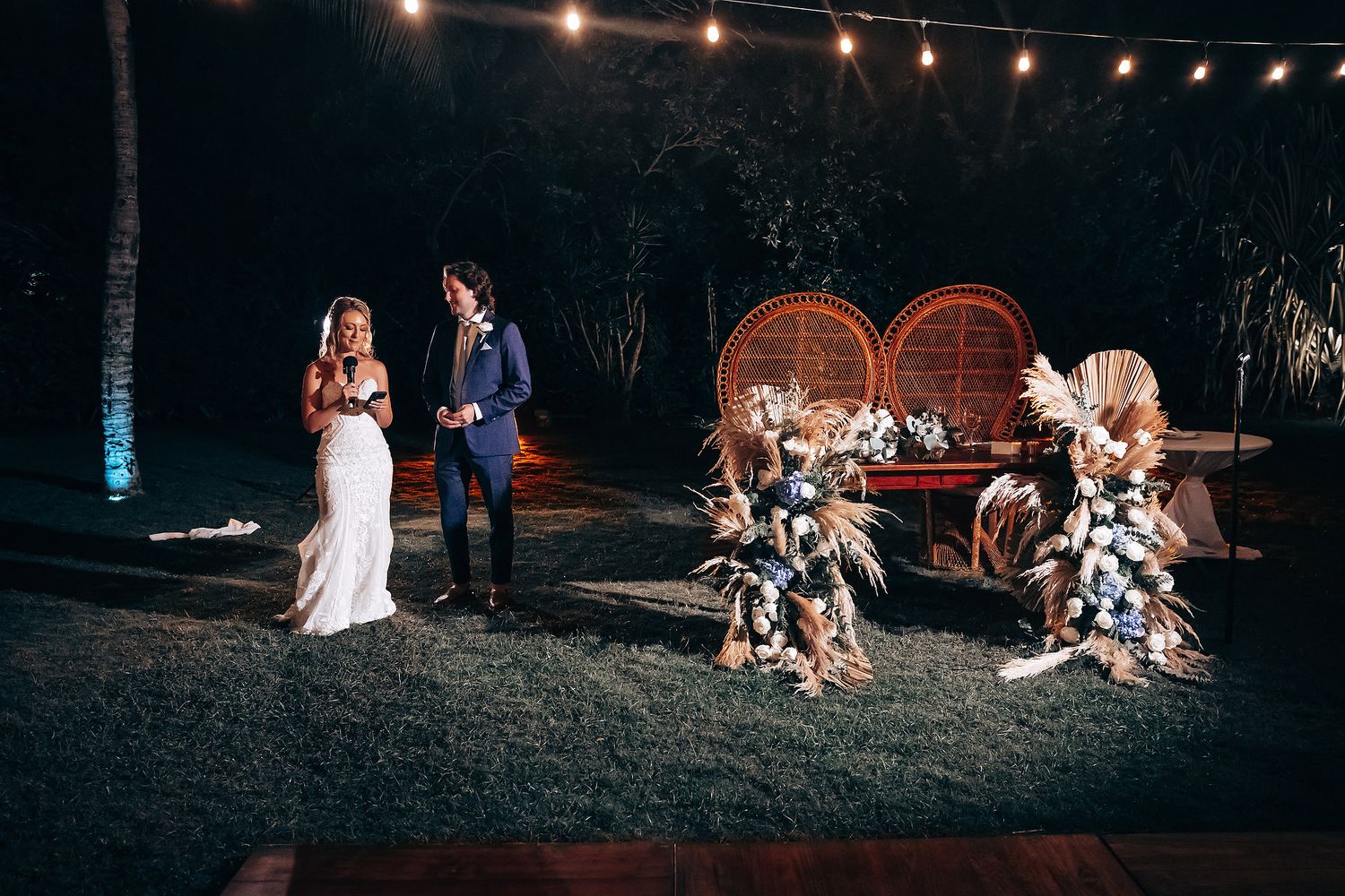 Outdoor wedding reception at night illuminated by warm string lights.