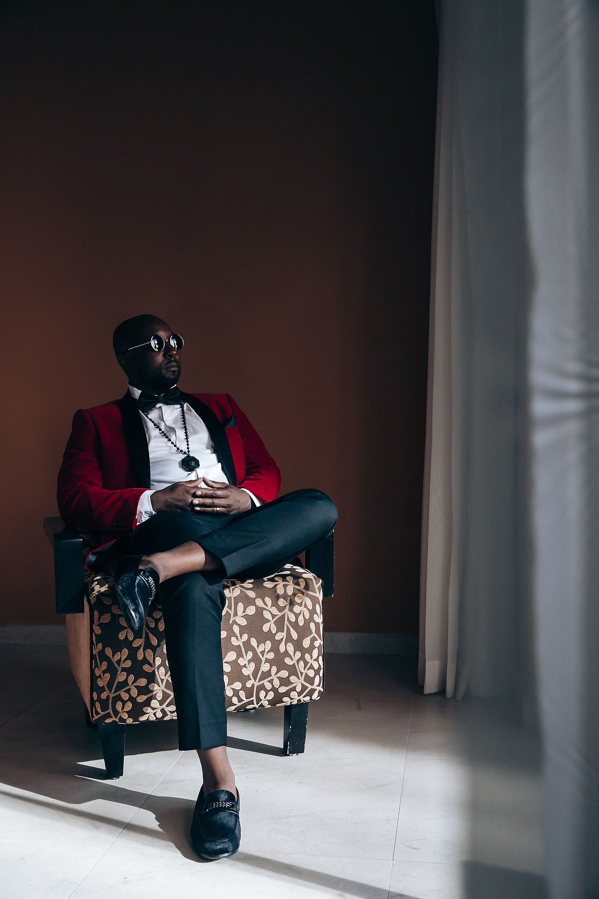Stylishly dressed person in red velvet blazer and sunglasses sitting on leopard print ottoman in moody lighting.