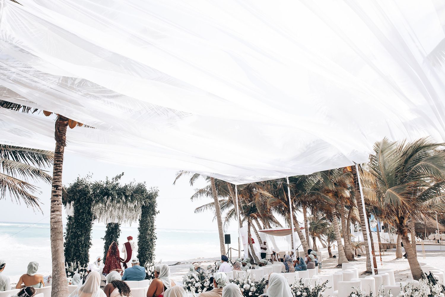 White fabric canopy stretches along a beach with palm trees and ocean backdrop.