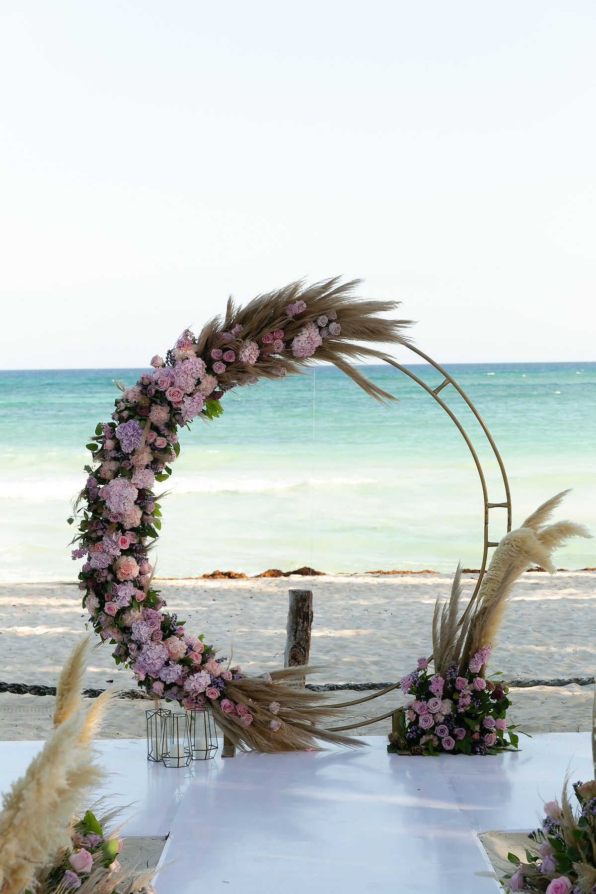 Curved floral wedding arch with pink and purple flowers and pampas grass stands against turquoise ocean waters.