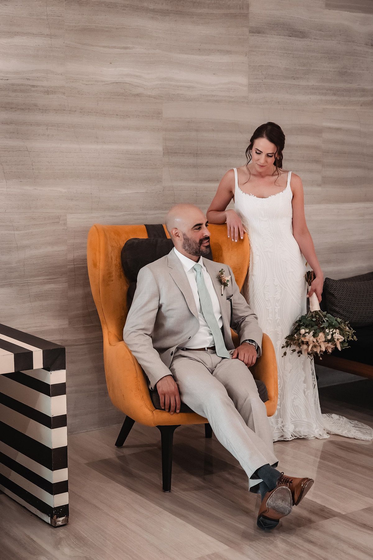 A wedding couple poses in a modern setting with an orange accent chair and striped decor elements.