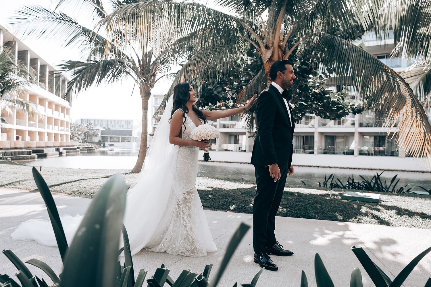 A wedding couple stands beneath swaying palm trees at a tropical beachfront resort venue.