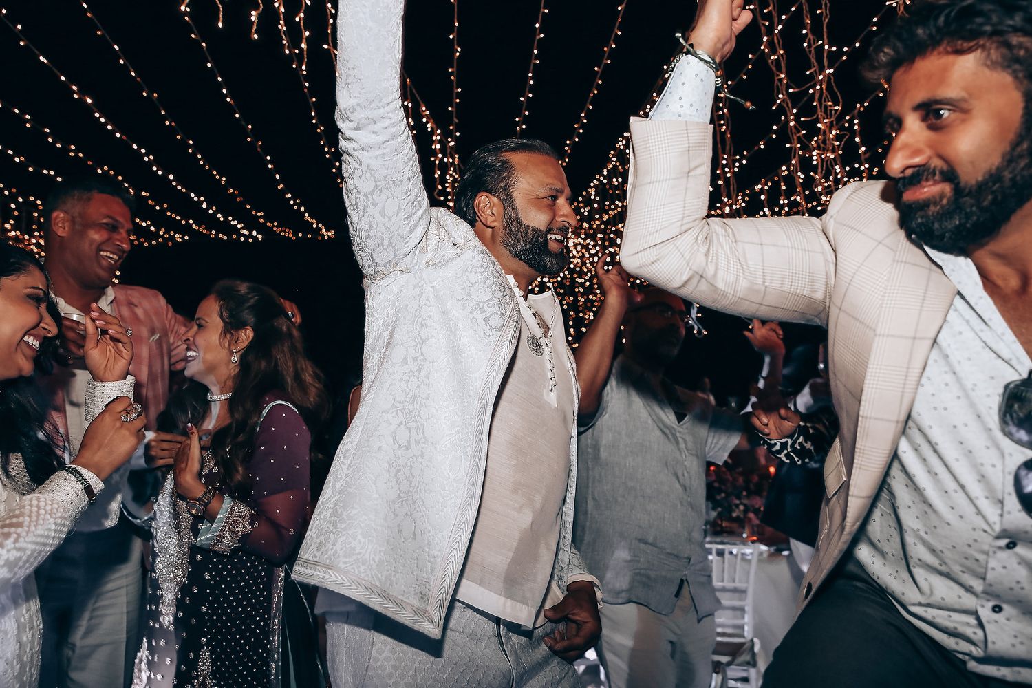 People dancing and celebrating at a lively nighttime party with string lights overhead and white attire.