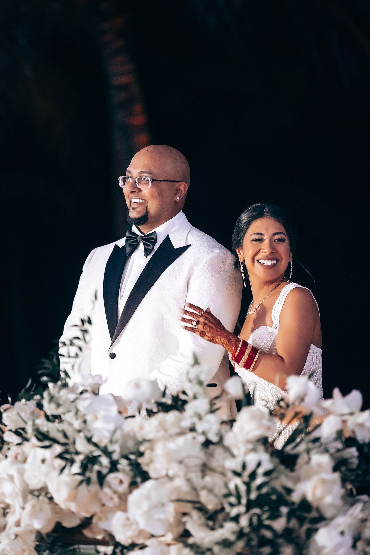 Wedding couple seated at reception table decorated with white floral arrangements.