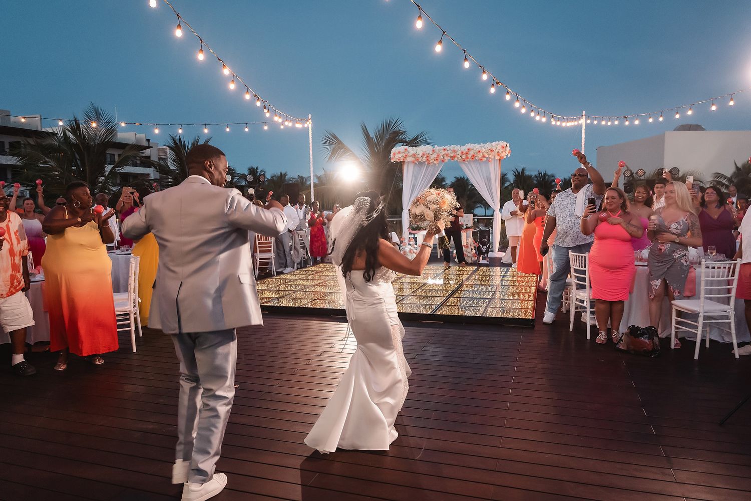 String lights illuminate an outdoor deck party setting at dusk with tables and chairs arranged for guests.