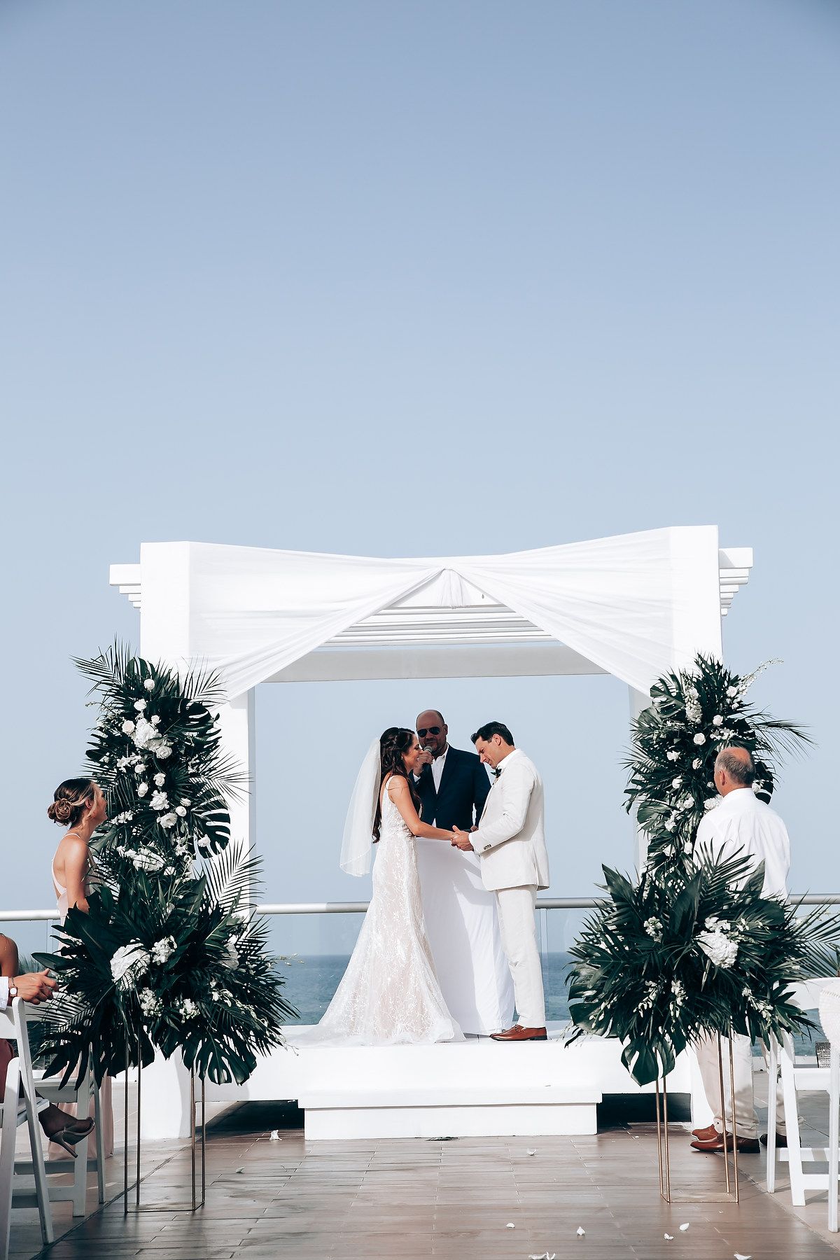 Beachside wedding ceremony with white gazebo and tropical palm arrangements under blue skies.
