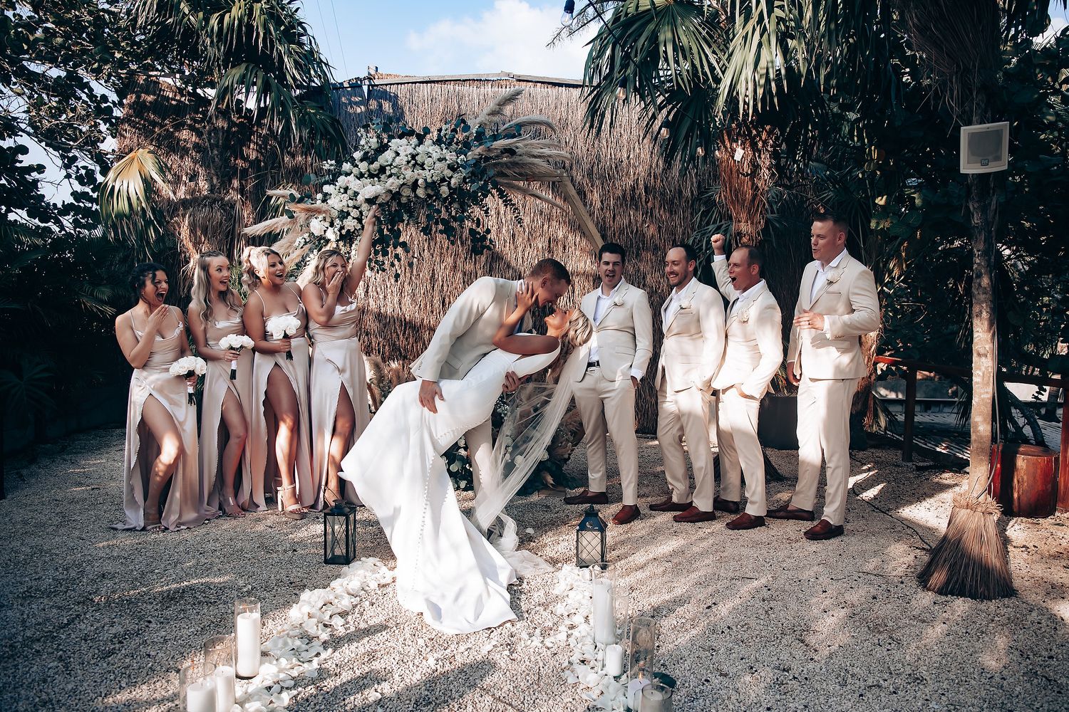 Wedding celebration in a tropical garden setting with guests seated on rustic benches.