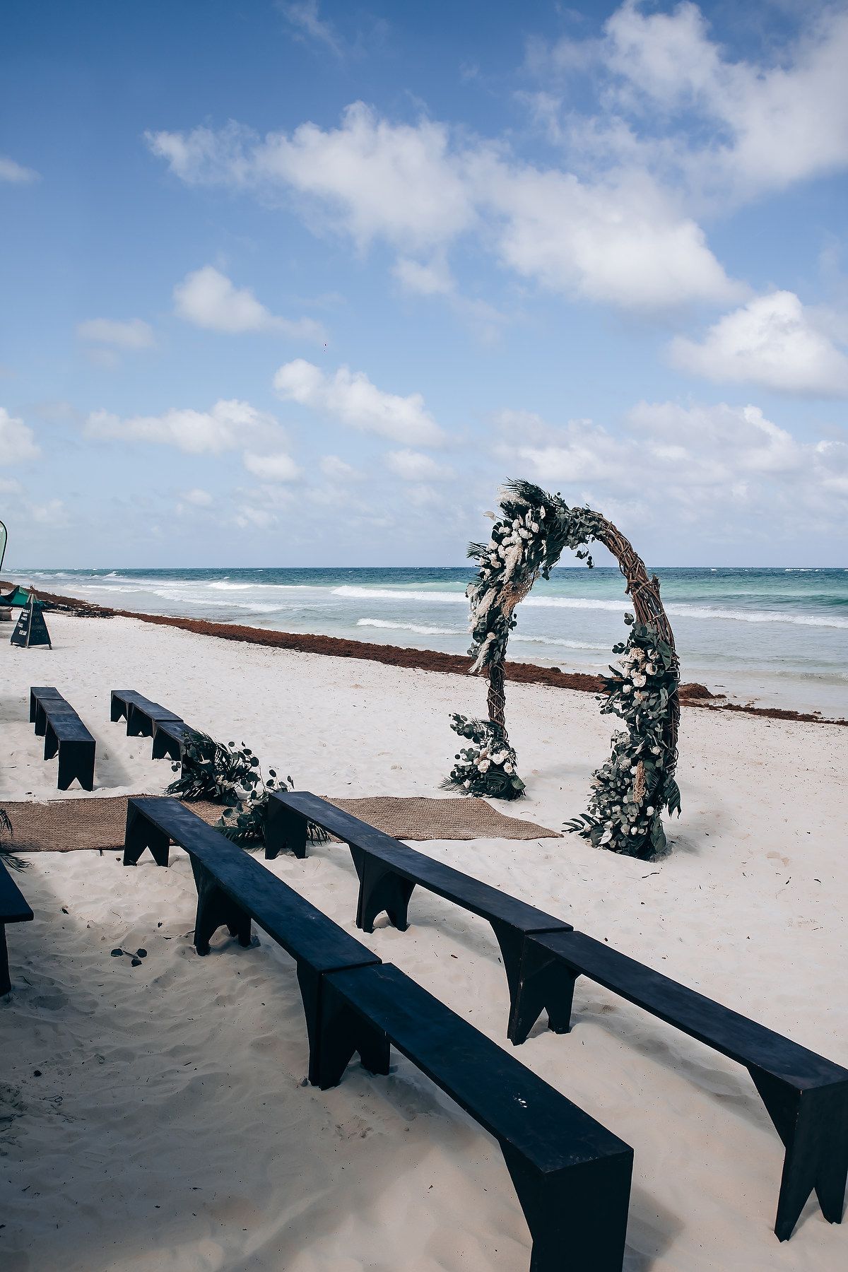 Dark wooden benches arranged for a beach wedding ceremony with floral arch and ocean view.