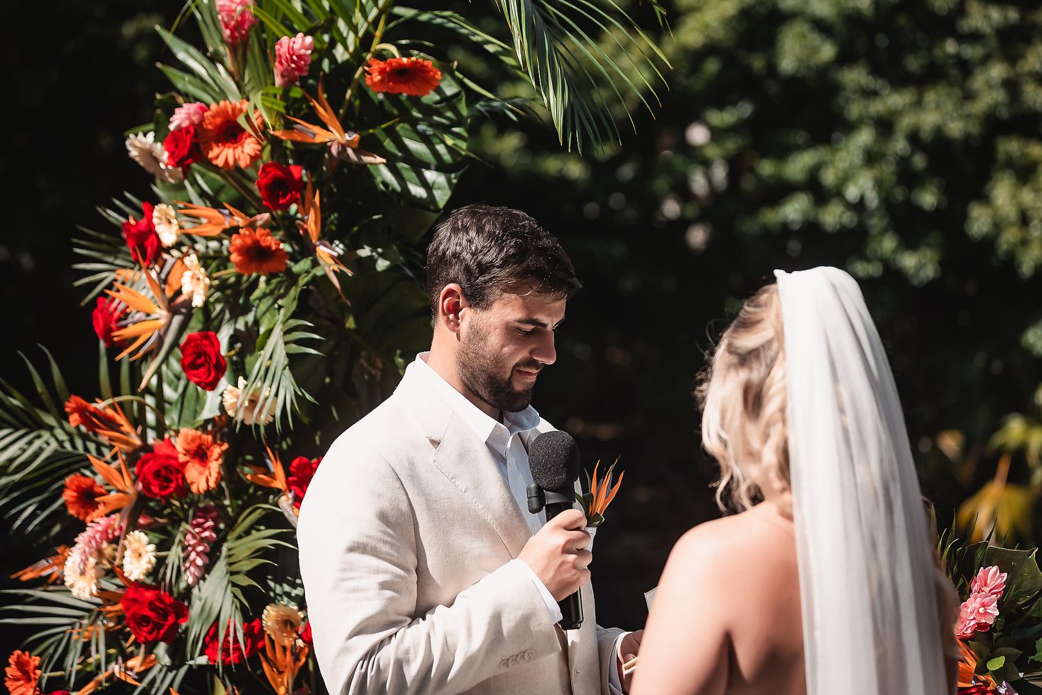 Tropical wedding ceremony with vibrant red floral arch and palm trees in the background.