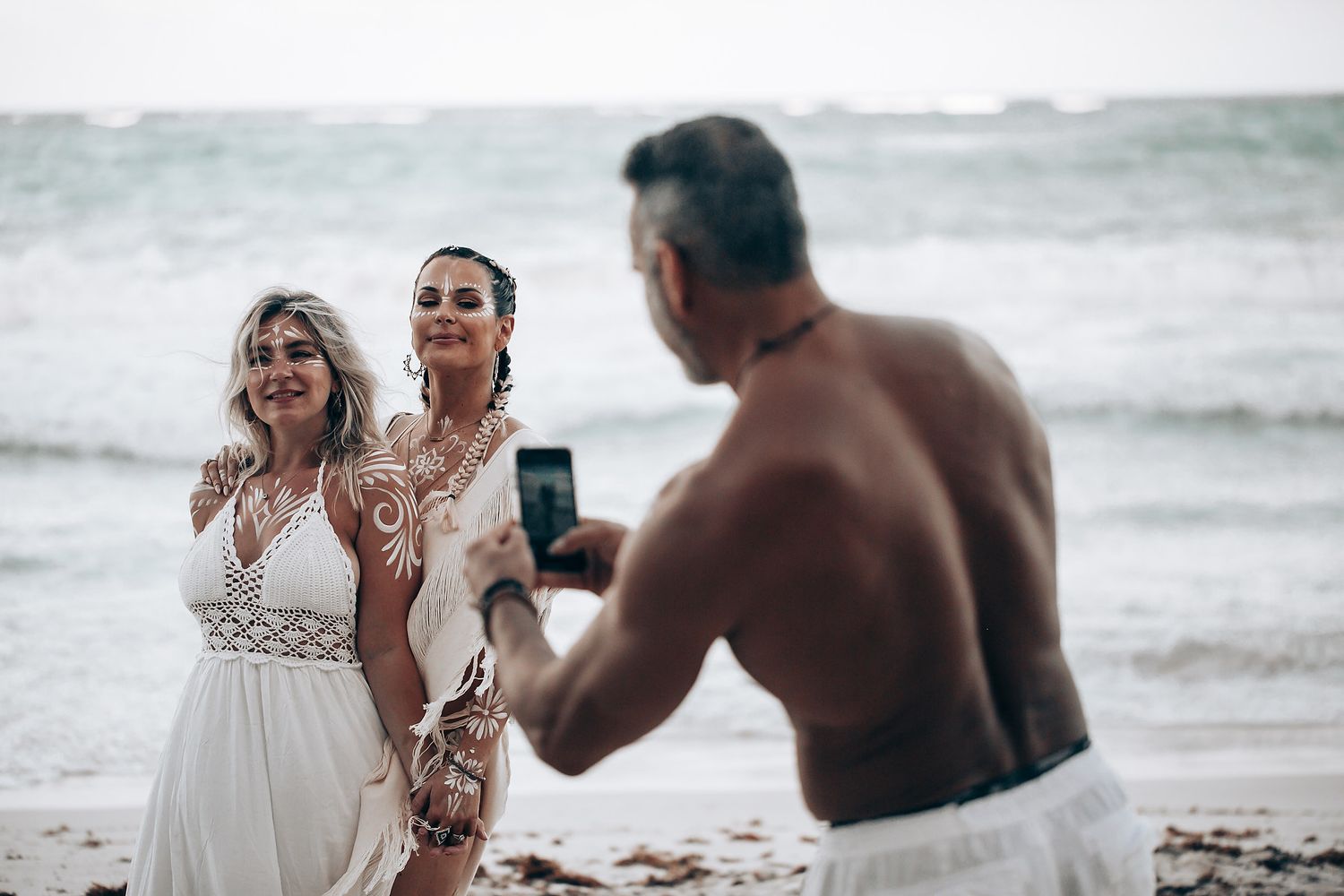 A beachside celebration with people in white attire posing for photos by the ocean.