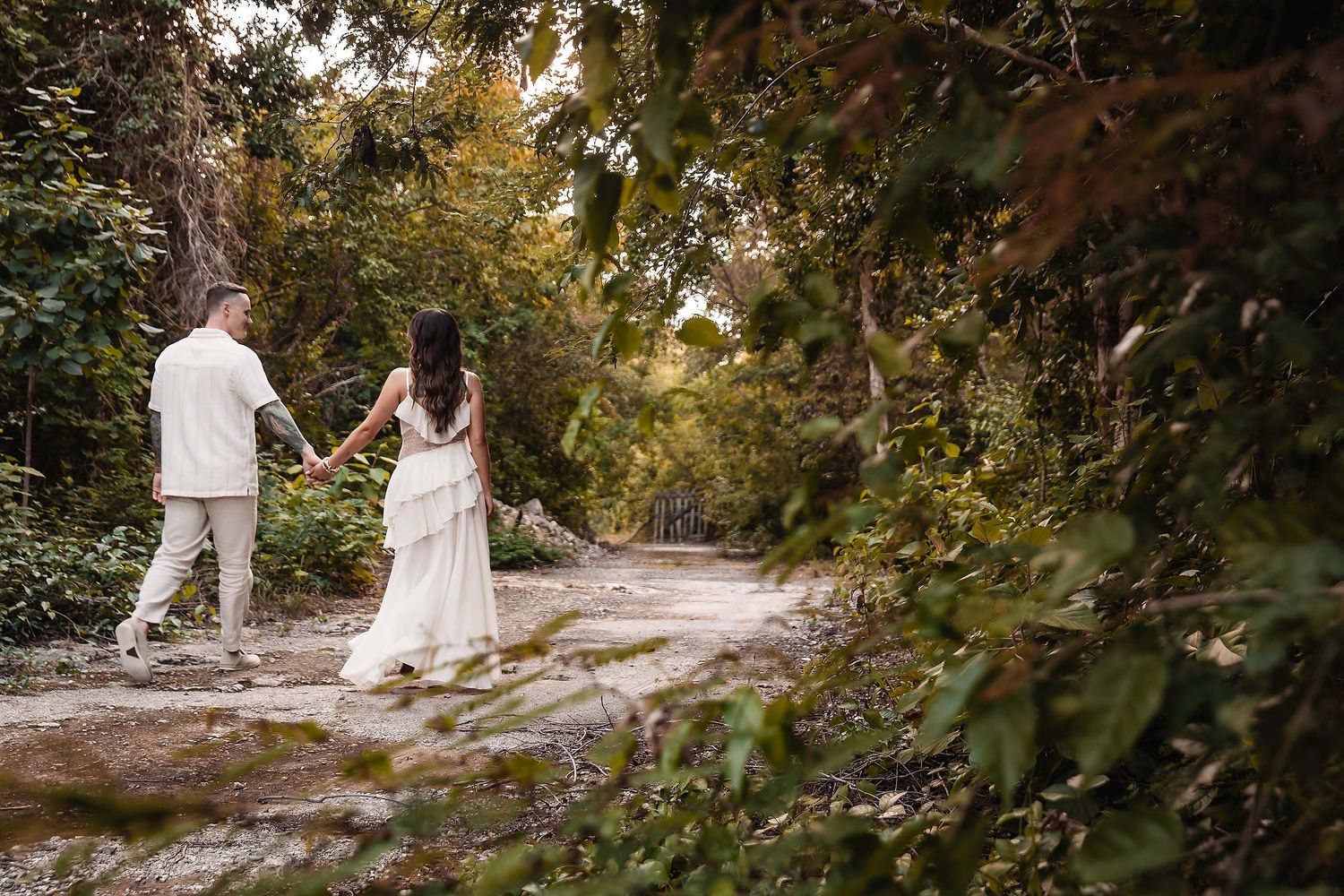 A couple in light colored outfits walks through a sunlit autumn forest path.