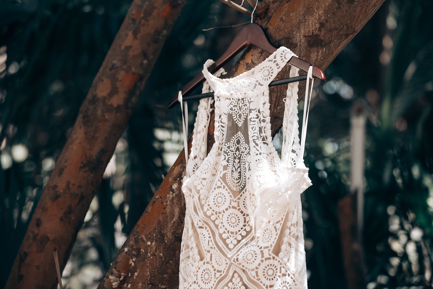 A delicate white lace wedding dress hangs from a rustic wooden beam in a forest setting.