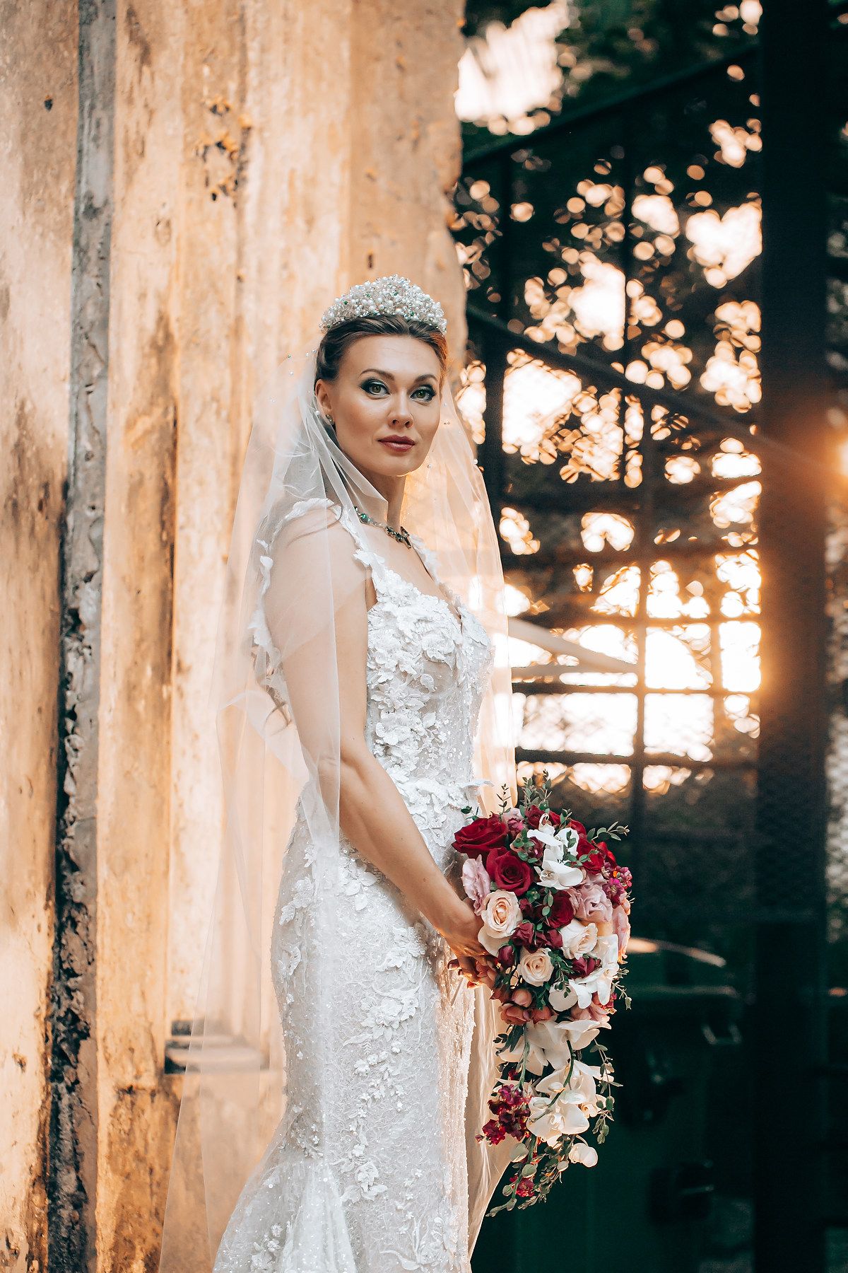 Elegant bride in lace dress holding pink and burgundy bouquet poses among sunlit trees at sunset.