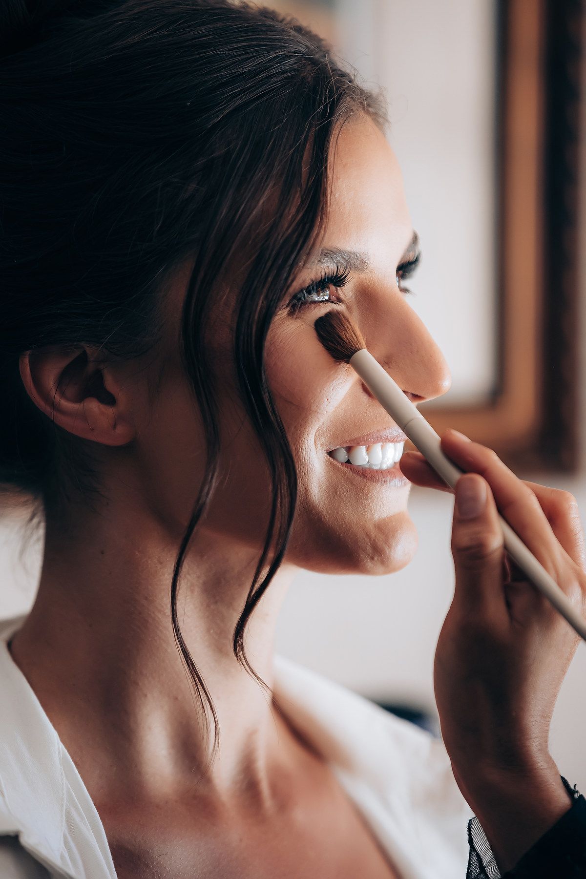 Makeup artist applies eyeshadow with a brush during bridal beauty preparation in natural window light.
