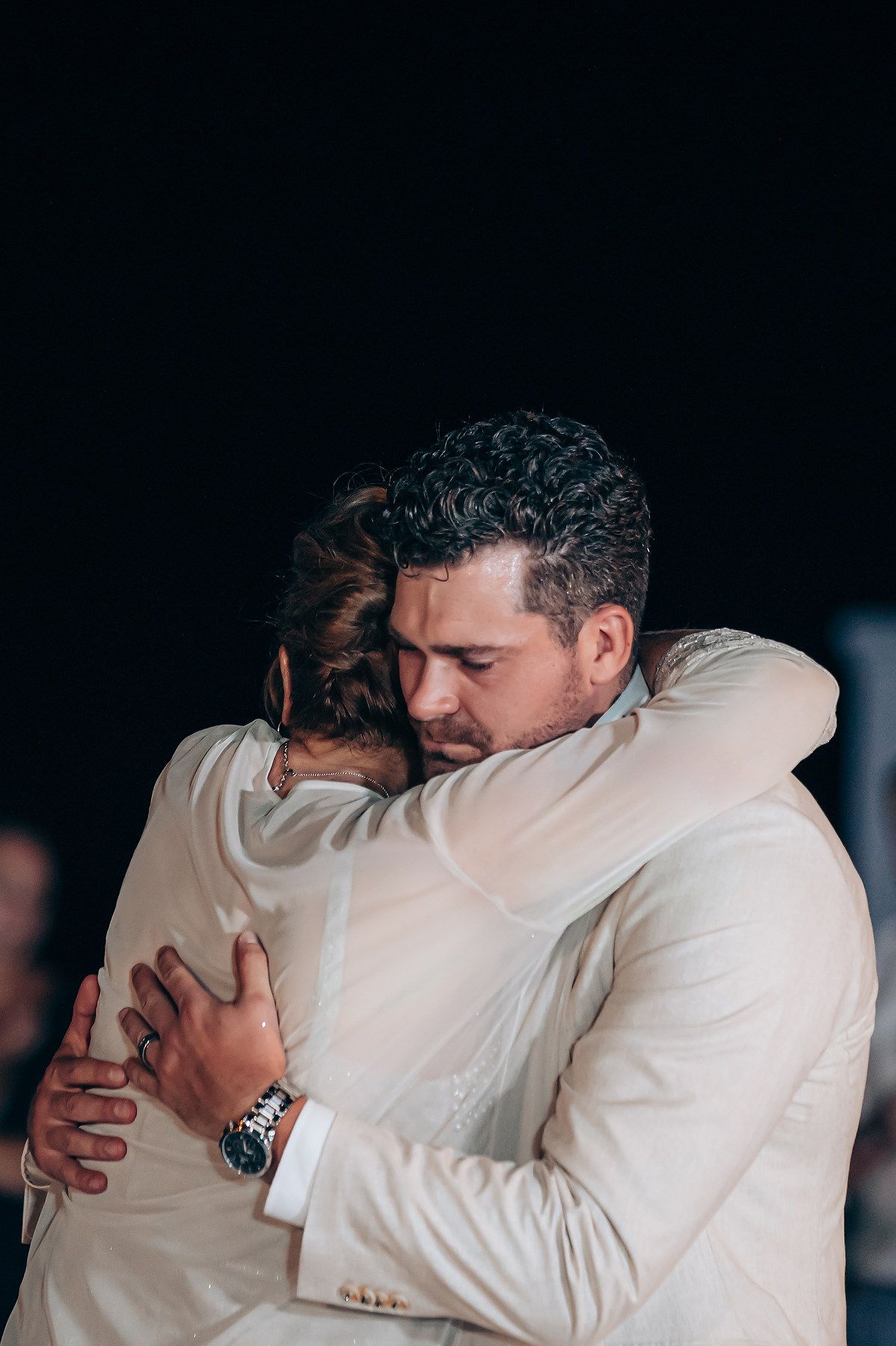 Two people in white attire share an emotional embrace against a dark background.