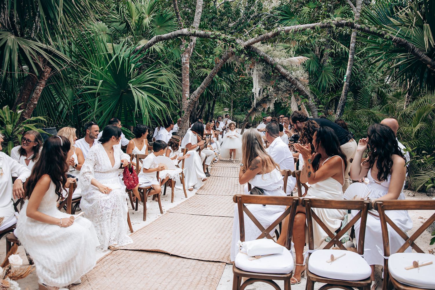 A panoramic view of a tropical beach wedding ceremony with guests dressed in white seated on wooden chairs.